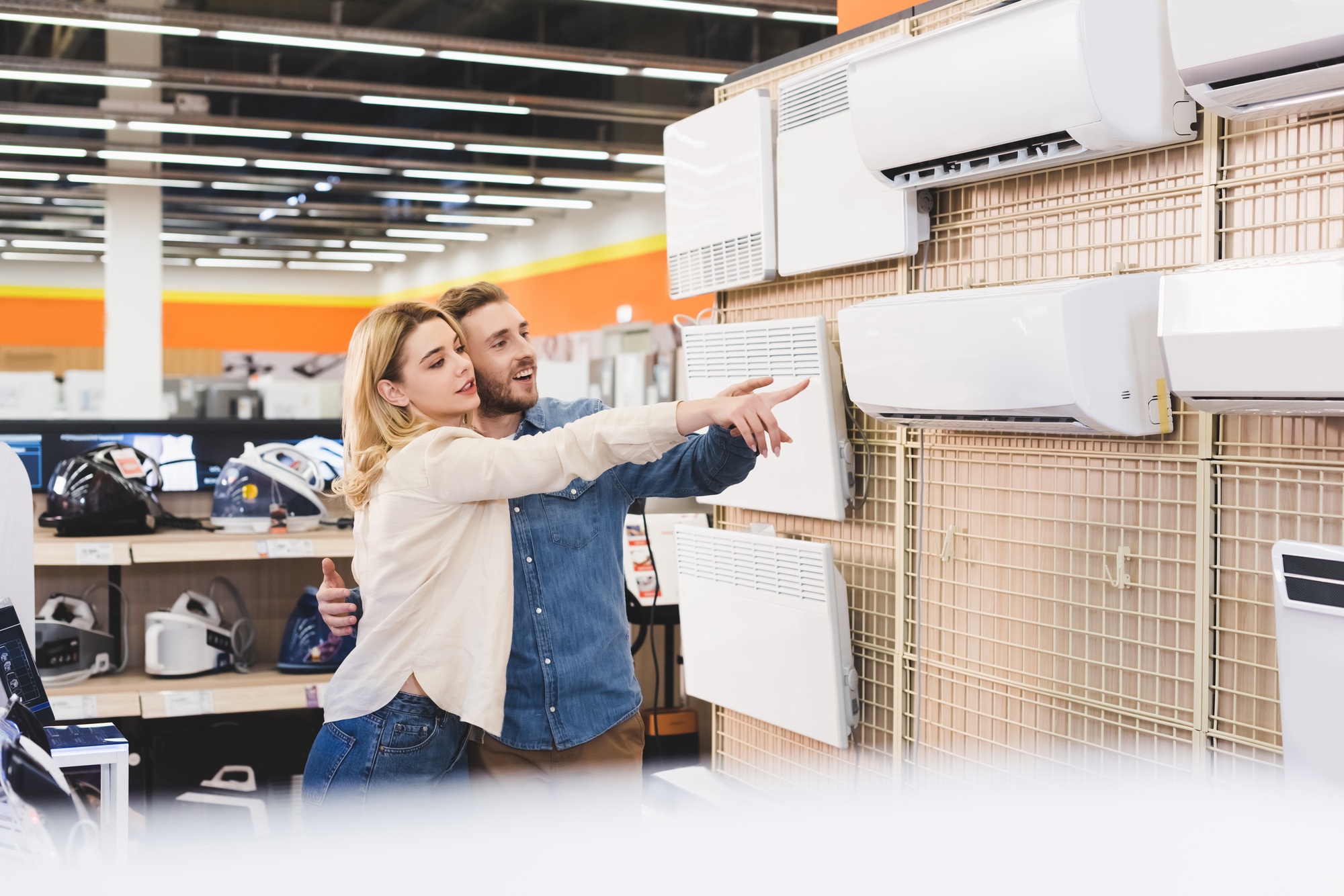 smiling boyfriend and girlfriend pointing with fingers at air conditioner in home appliance store